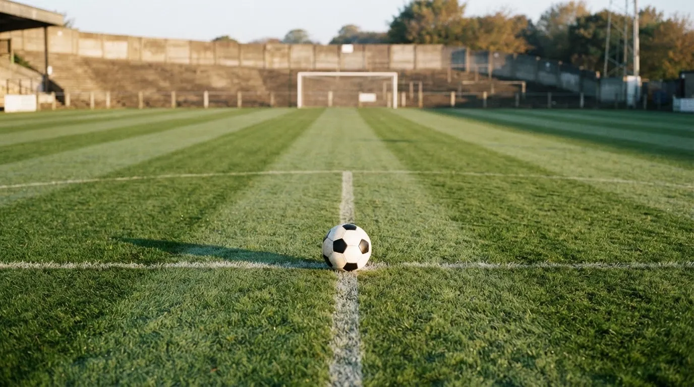Ein Fussball liegt auf dem Anstosspunkt eines leeren Rasenplatzes im frühen Morgenlicht, dahinter erhebt sich eine menschenleere Tribüne.