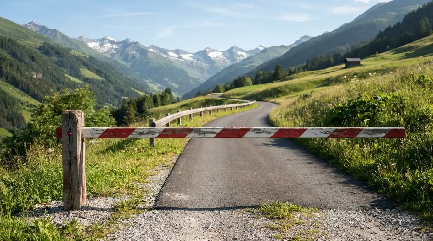 Ein Schlagbaum sperrt eine Landstrasse in den Bergen, dahinter windet sich der Weg zwischen grünen Hängen.