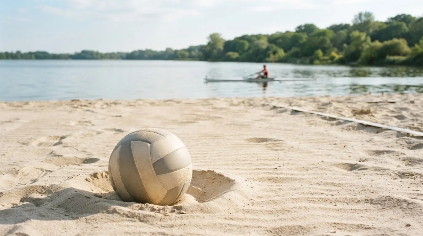 Ein Volleyball liegt im Sand eines Spielfelds am Seeufer, im Hintergrund gleitet ein Boot über das Wasser.