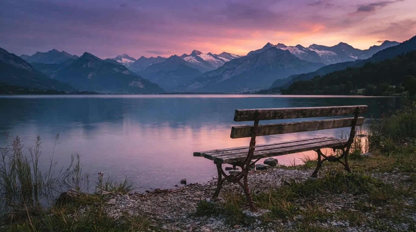 Eine Holzbank steht in der Dämmerung am Seeufer, im Hintergrund zeichnet sich ein Bergpanorama gegen den violetten Himmel ab.