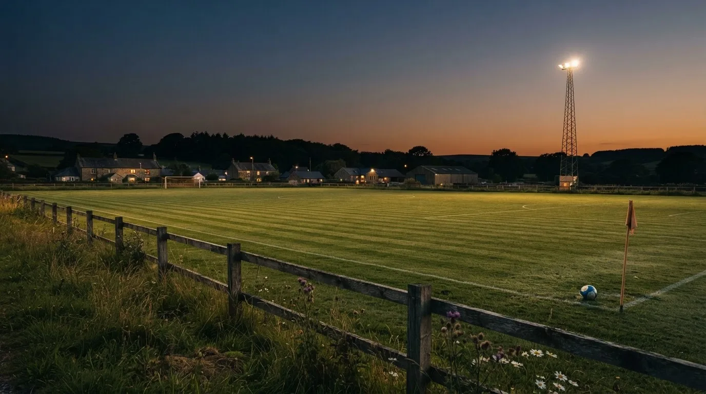 Ein kleiner Dorf-Fussballplatz liegt in der Dämmerung unter einem hohen Flutlichtmast, am Spielfeldrand steht ein Holzzaun.