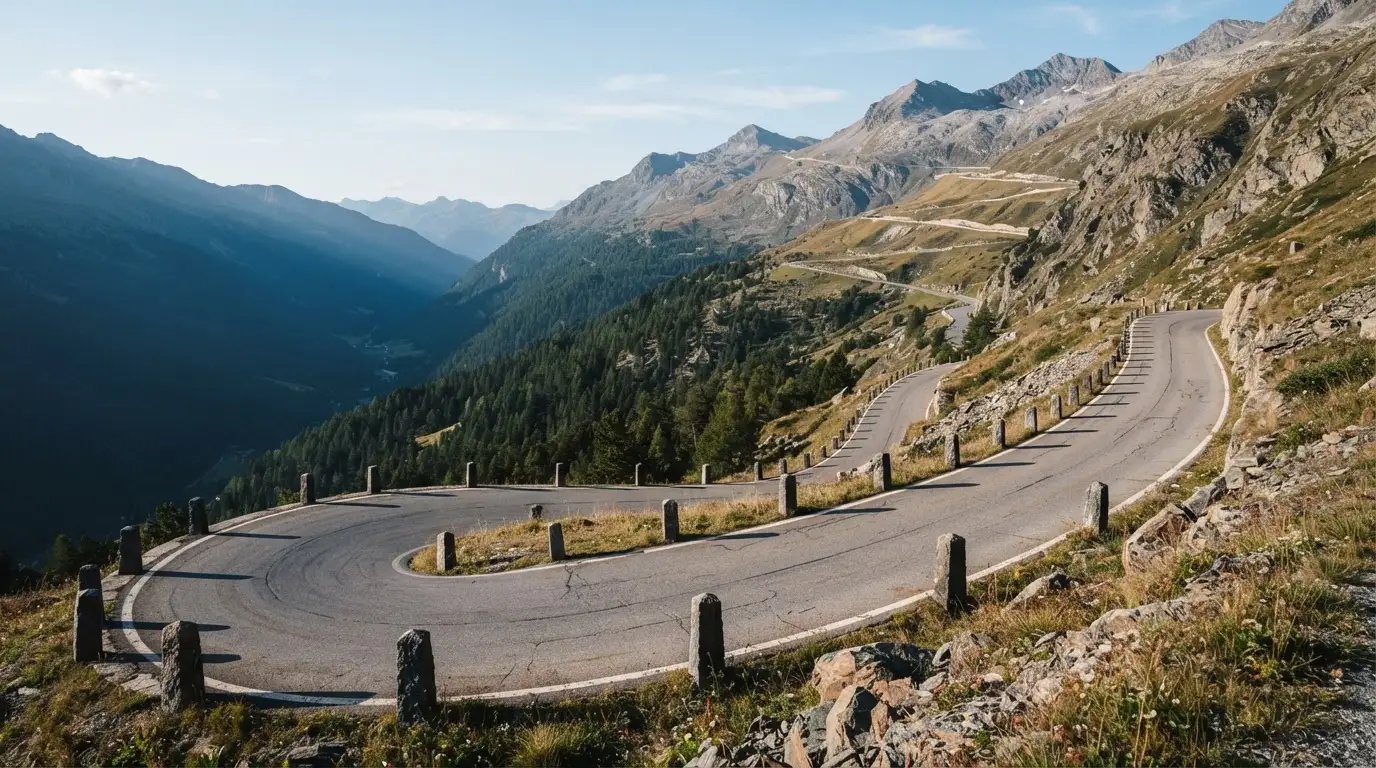 Eine leere Passstrasse mit Serpentinen windet sich im Morgenlicht einen Berg hinauf, am Rand stehen niedrige Steinpfosten.