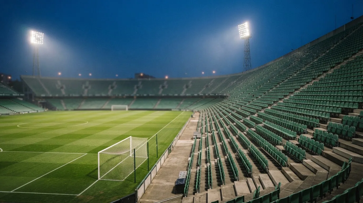 Leere Tribünenränge eines Fussballstadions in der Abenddämmerung, über dem Rasen leuchten die Flutlichter.