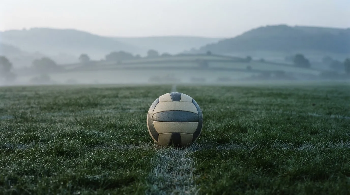 Ein Fussball liegt am frühen Morgen auf dem Anstosspunkt eines taunassen Rasens, im Hintergrund erheben sich Hügel im Dunst.