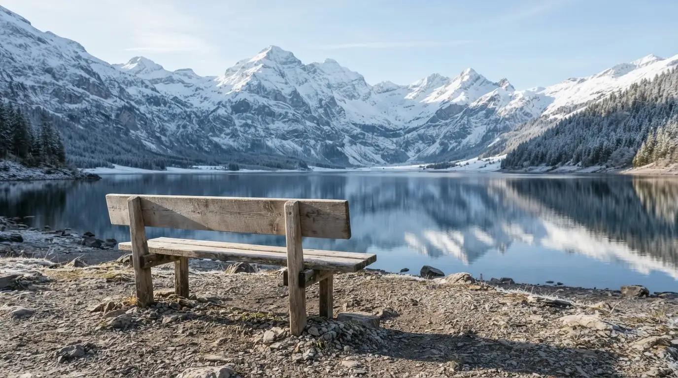 Eine Bank an einem Bergsee, dahinter schneebedeckte Gipfel im klaren Licht eines Wintermorgens.