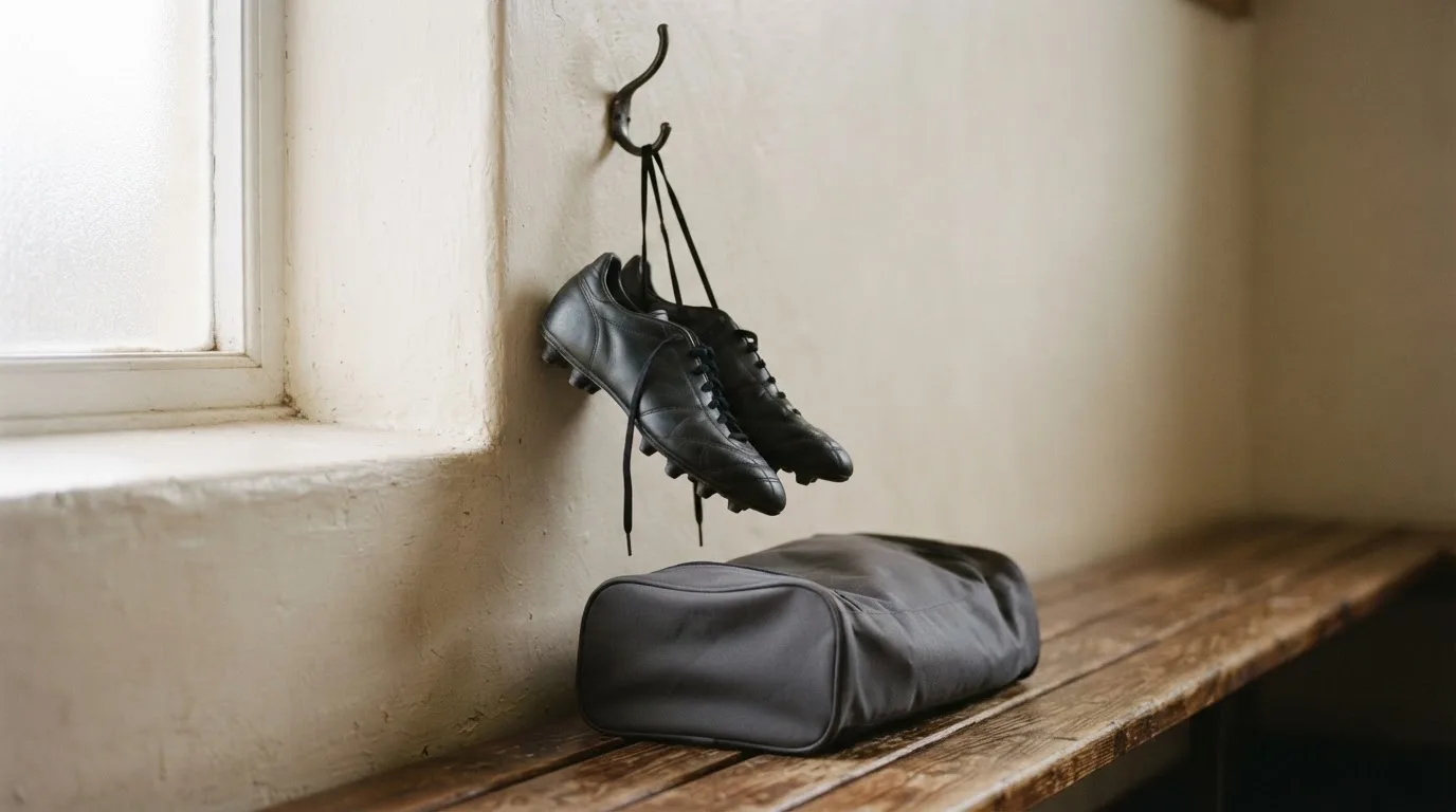 Ein Paar Fussballschuhe hängt an einem Haken in einer ruhigen Umkleide, darunter steht eine Sporttasche auf einer Holzbank.