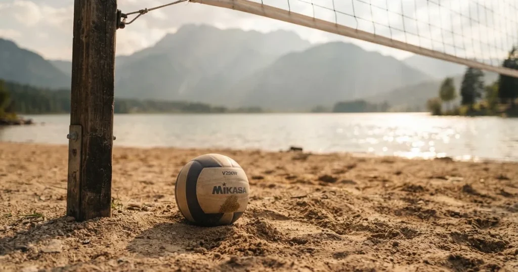 Ein Volleyball liegt am Fuss des Netzes auf Sand, im Hintergrund glitzert ein See und ein Bergzug steht im Dunst.