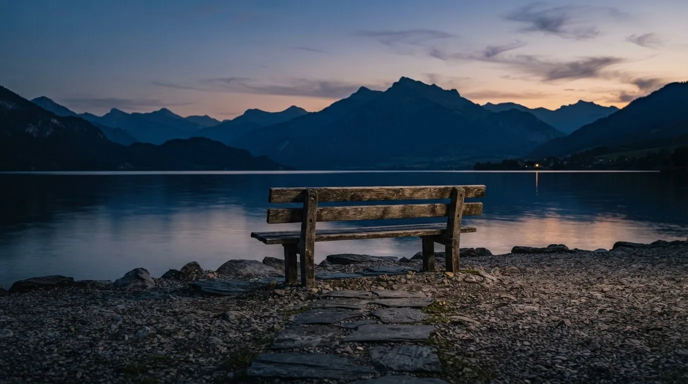 Eine leere Holzbank am Seeufer in der Dämmerung, im Hintergrund dunkle Berge