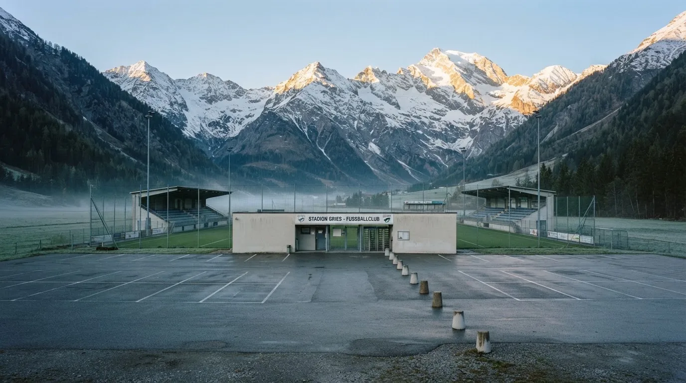 Schneebedeckte Berge ragen hinter einem kleinen Stadion in einem Alpental auf, im Vordergrund ein leerer Parkplatz am Morgen.