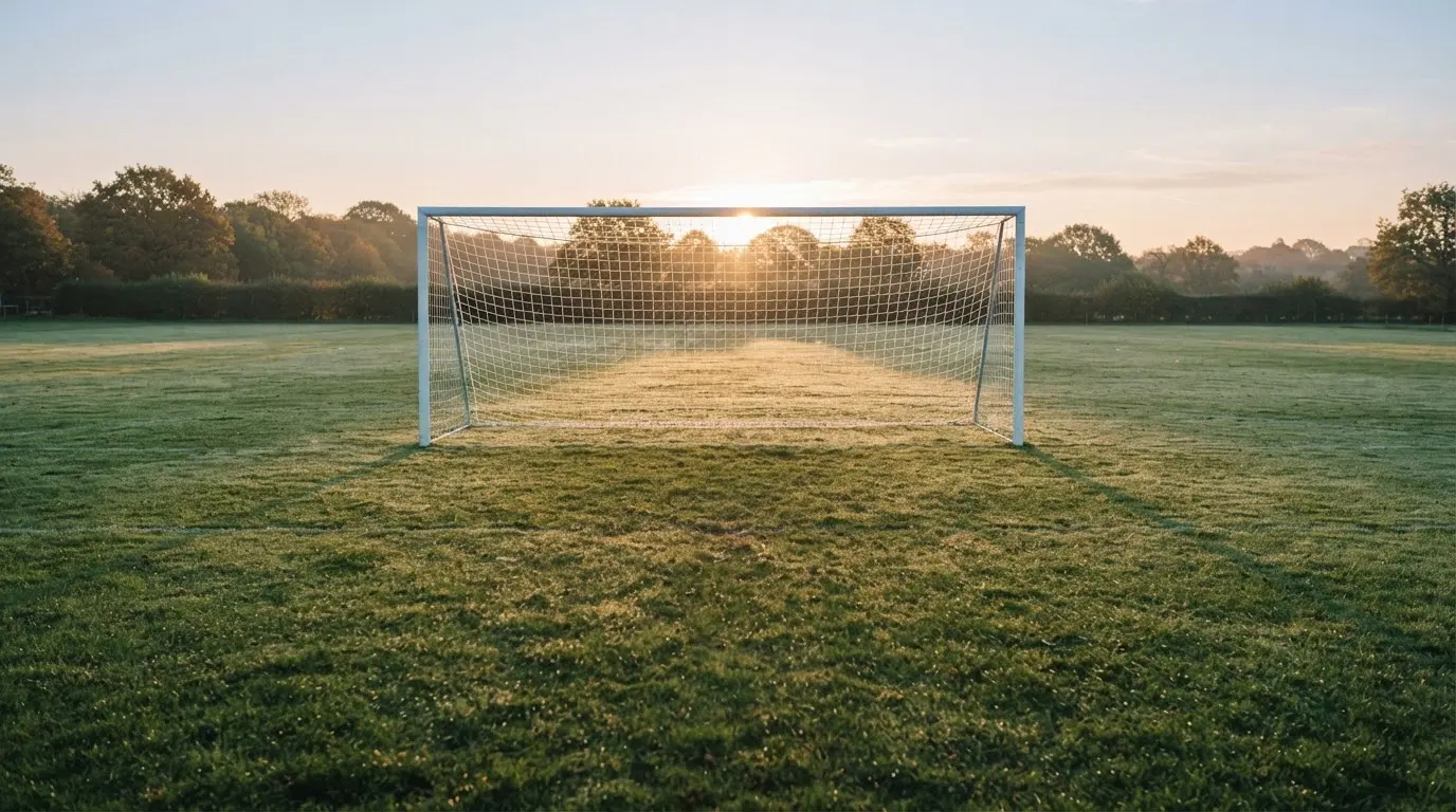 Ein Fussballtor mit Netz in der Totalen auf einem leeren Platz, Tau glänzt im Gras und die Morgensonne steht tief.