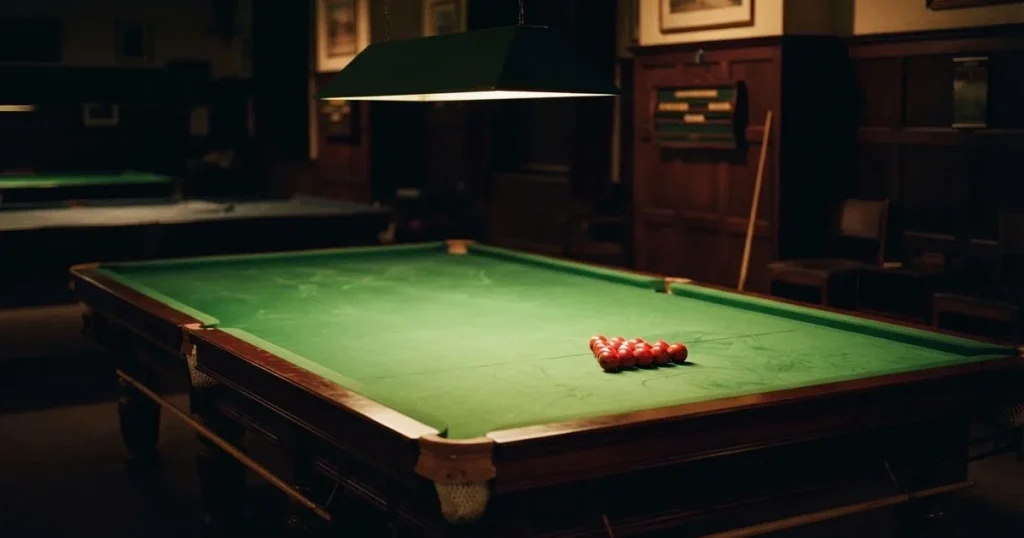 Ein Snookertisch mit grünem Tuch und einem Dreieck aus roten Kugeln unter einer tief hängenden Lampe in einer leeren Halle.