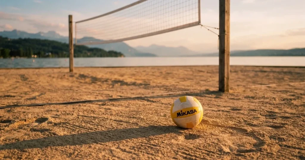 Ein Volleyball liegt im Sand eines Beachplatzes, zwischen zwei Pfosten ist ein Netz gespannt, tiefe Abendsonne wirft lange Schatten.