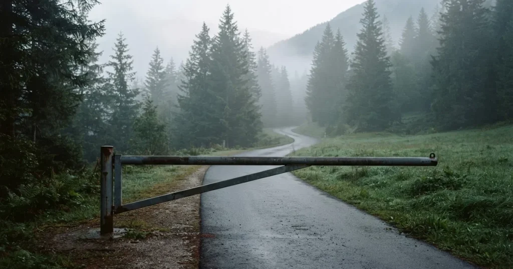 Eine geschlossene Schranke steht an einer ruhigen Bergstrasse im Morgennebel, dahinter Tannen und eine nasse Wiese.