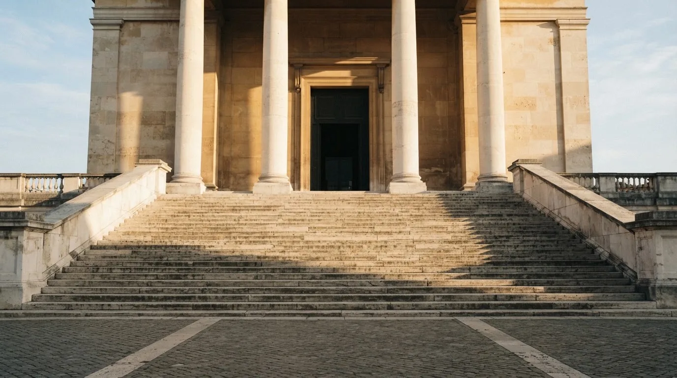 Eine steinerne Treppe führt zu einem Portal mit Säulen im Morgenlicht, der Platz davor ist leer.
