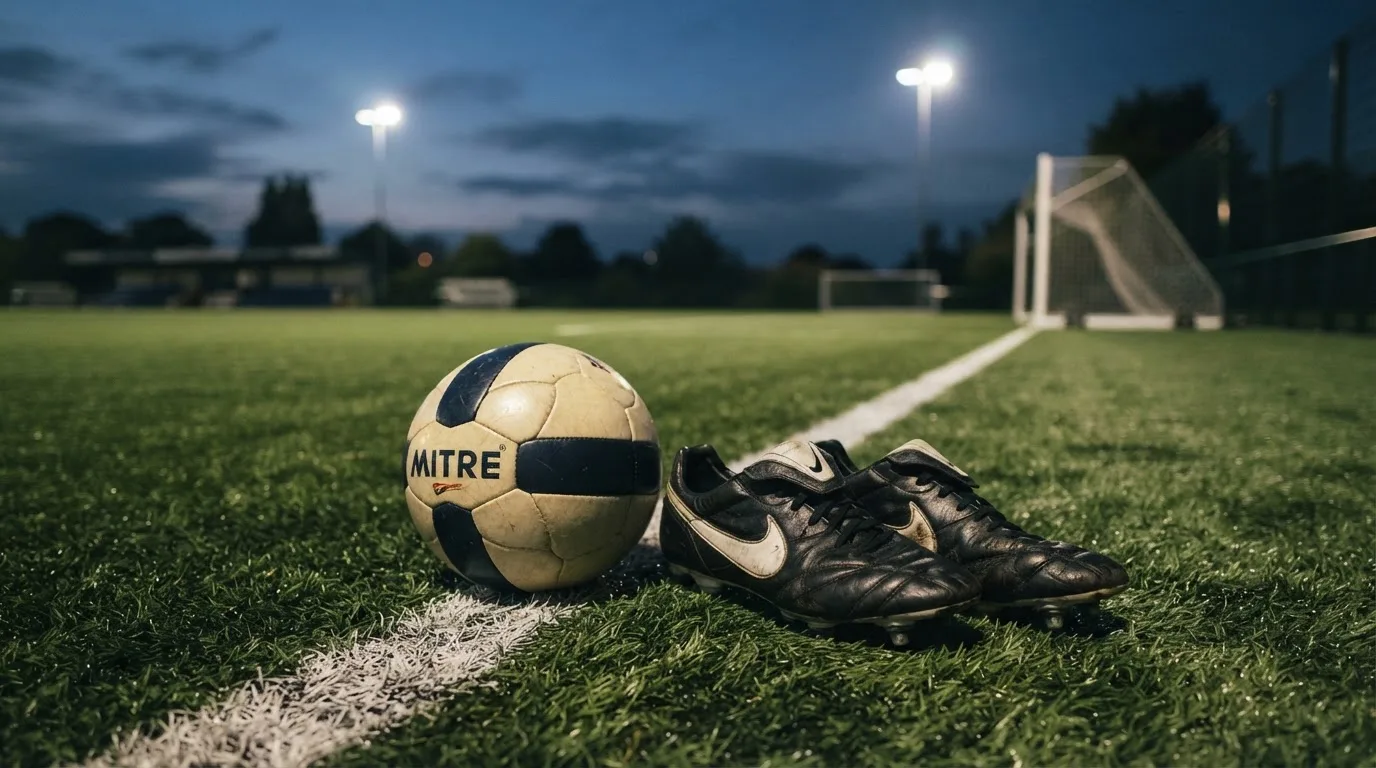 Ein Fussball und ein Paar Stollenschuhe stehen am Rand eines Kunstrasenplatzes, Flutlichtmasten ragen in den Abendhimmel.