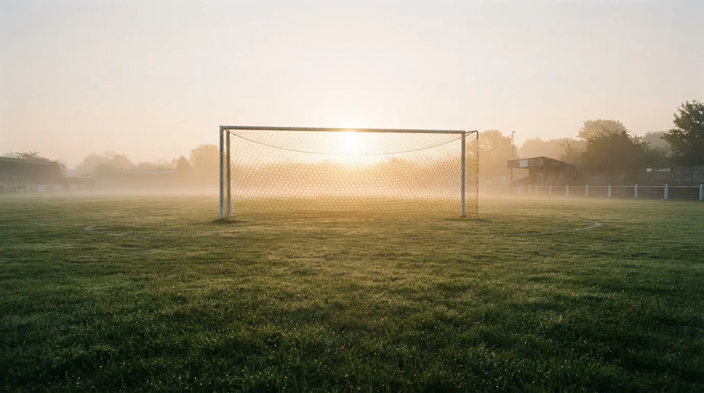 Ein Torpfosten mit Netz steht im Gegenlicht auf einem leeren Fussballplatz, über dem Rasen liegt Nebel.