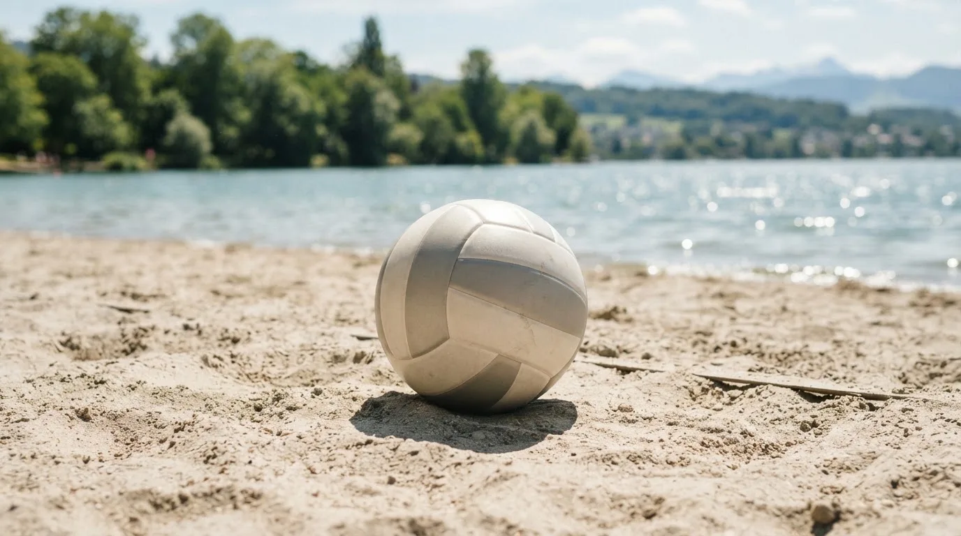 Ein Volleyball liegt auf dem Sand eines Strandfeldes am Seeufer, im Hintergrund glitzert das Wasser.