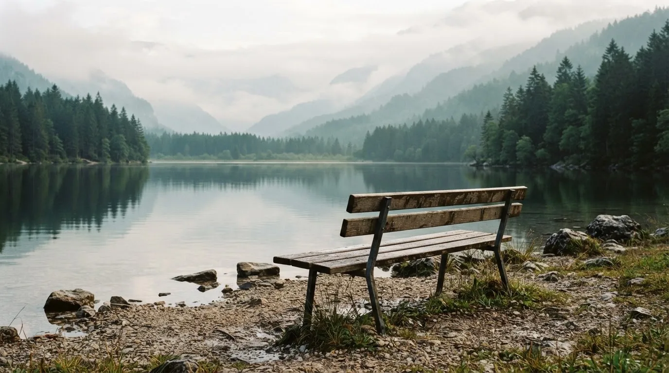 Eine Holzbank am Ufer eines ruhigen Bergsees, am gegenüberliegenden Ufer steigen dunkle Hänge in den Frühnebel