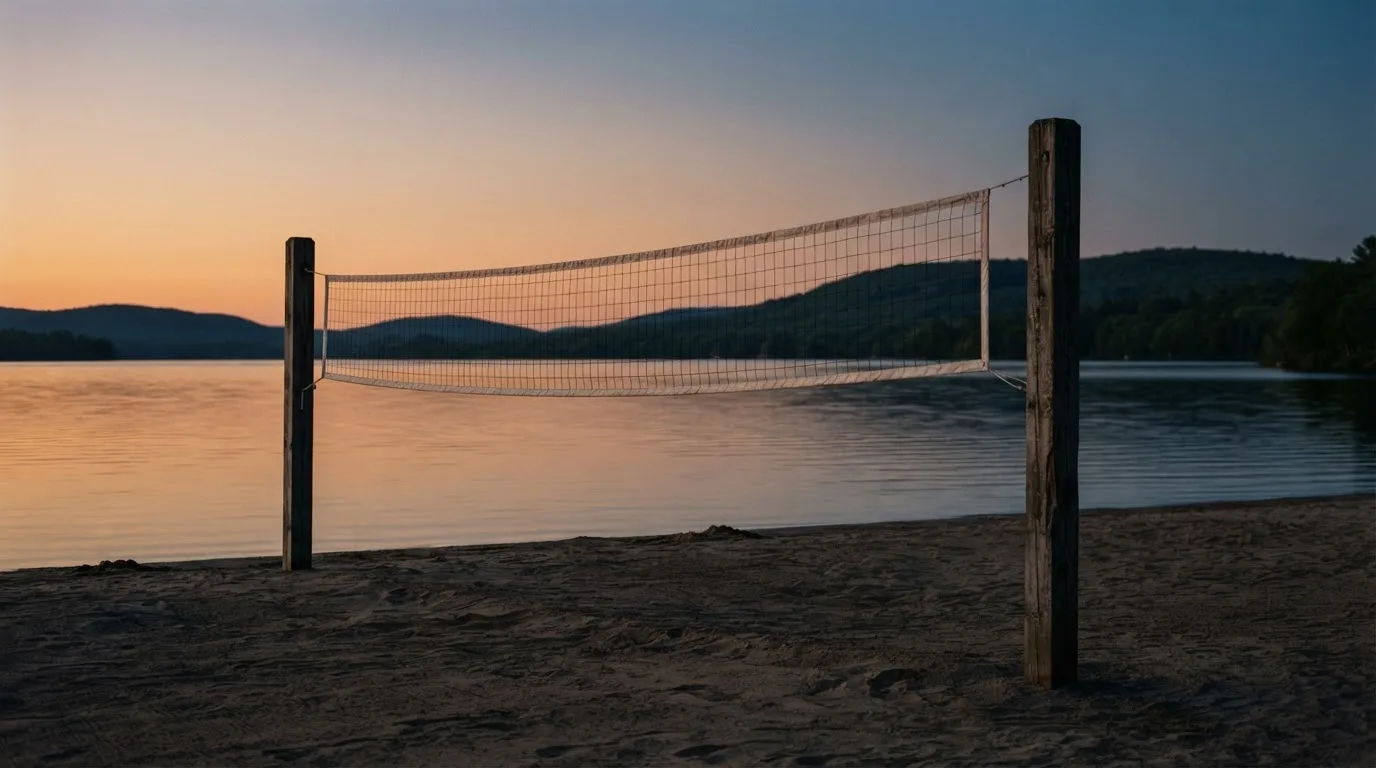Ein Volleyballnetz steht an einem Seeufer in der Abenddämmerung, der Sand ist leer und das Wasser glatt.