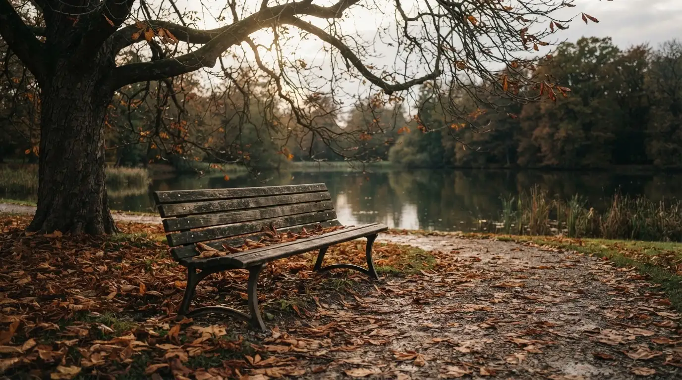 Eine Parkbank unter einer Kastanie, Herbstlaub bedeckt den Kiesweg, im Hintergrund ein stiller Teich.