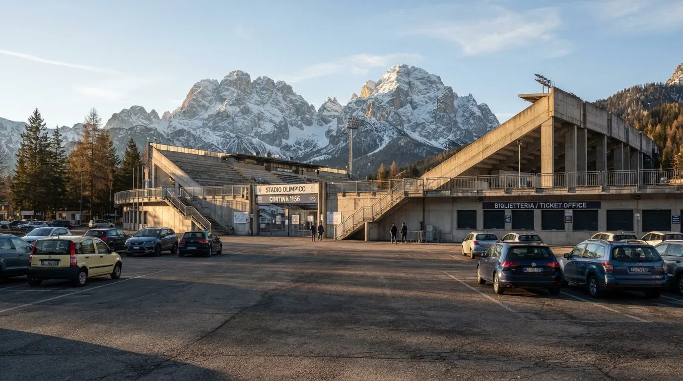 Hinter einem leeren Stadion ragen verschneite Berggipfel in den klaren Morgenhimmel, davor ein Parkplatz im ersten Sonnenlicht.