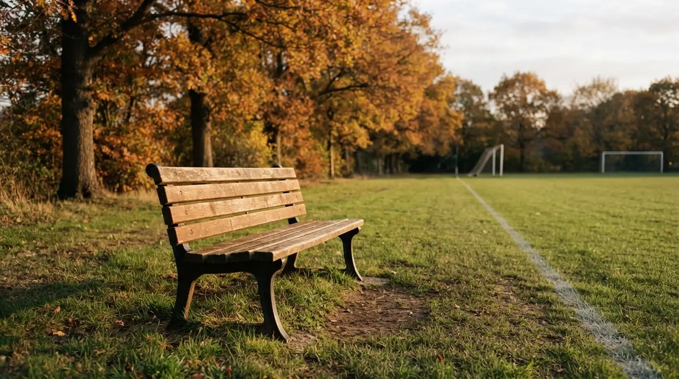 Eine hölzerne Bank steht an der Seitenlinie eines leeren Sportplatzes, dahinter Bäume im Herbstlicht.